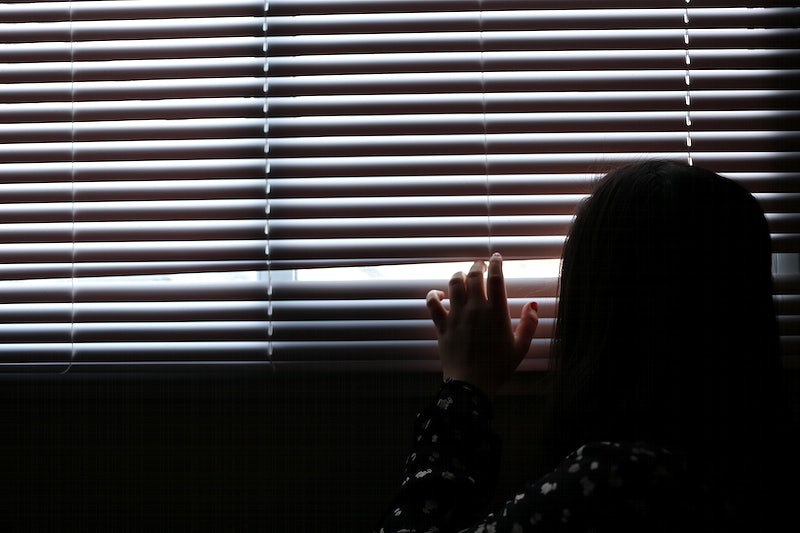 A girl peaks through window blinds out of a dark room.